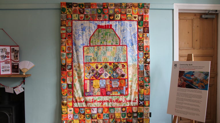 A colourful patchwork quilt featuring pockets representations of what home means hangs from the wall.